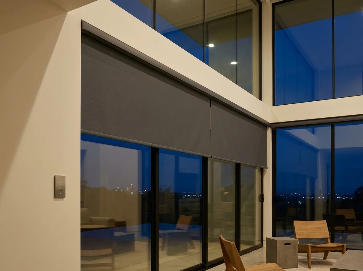 Motorised blind spanning a tall double-volume glass wall in a Baronetcy Estate home, wall-mounted control panel visible, city lights beyond at dusk